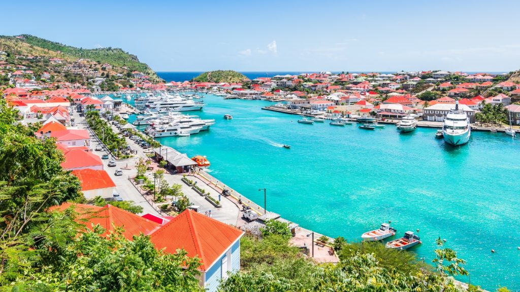 Aerial view of the vibrant harbor in Saint Barthelemy/St. Barths, featuring turquoise waters, red-roofed waterfront buildings, and a bright blue sky.