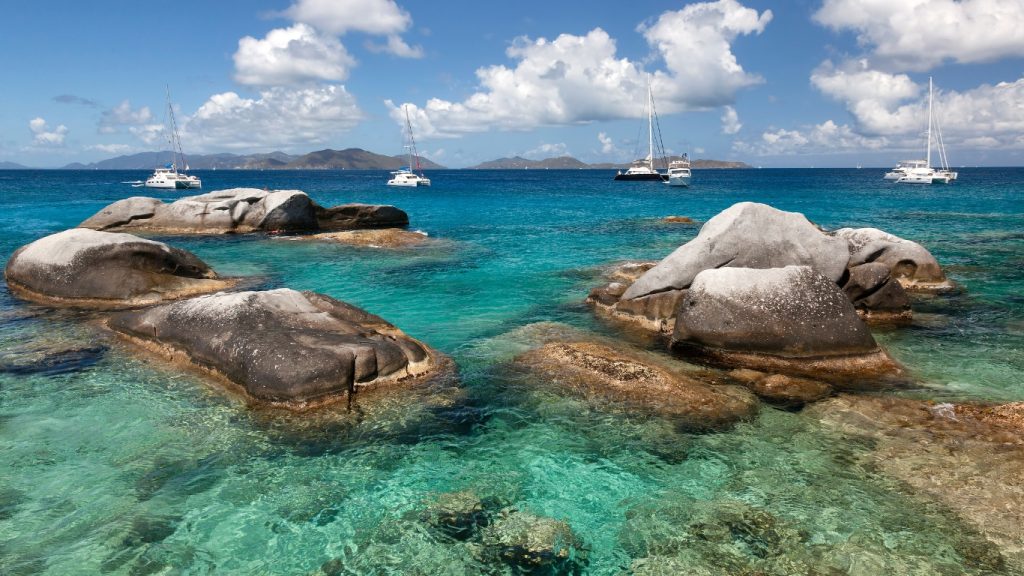 Giant granite boulders dot the turquoise waters at The Baths on Virgin Gorda, British Virgin Islands, with boats moored in the distance.